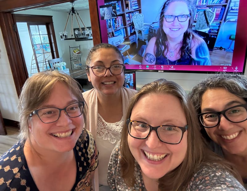 Four women smile into the camera. A fifth woman on a screen behind them also smiles into the camera